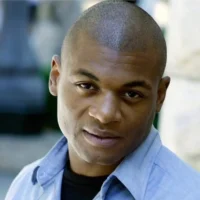 A man with short hair and a light blue collared shirt looks directly at the camera with a neutral expression.