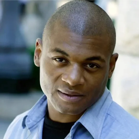 A man with short hair and a light blue collared shirt looks directly at the camera with a neutral expression.