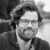 A man with wavy hair, glasses, and a beard wearing a collared shirt, smiling outdoors with a blurred background.