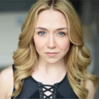 A woman with long blonde hair and blue eyes looks directly at the camera, wearing a black top with crisscross detailing at the neckline.