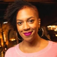 A woman with an asymmetrical hairstyle, hoop earrings, and bright pink lipstick smiles at the camera, wearing a light pink top in a warmly lit indoor setting.