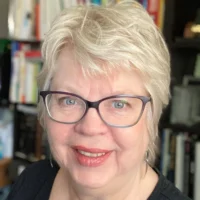 A woman with short gray hair and glasses smiles at the camera; bookshelves are visible in the background.
