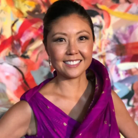 A woman wearing a purple sari smiles at the camera, standing in front of a colorful abstract background.