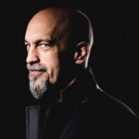 A bald man with a goatee and mustache, wearing a dark coat, is looking at the camera against a black background.