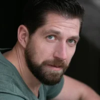 A bearded man with short dark hair and light eyes, wearing a green shirt, looks directly at the camera against a dark background.