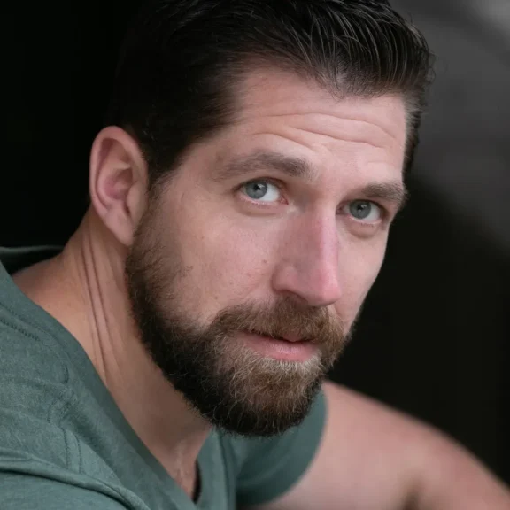 A bearded man with short dark hair and light eyes, wearing a green shirt, looks directly at the camera against a dark background.