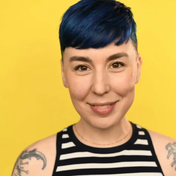 A person with short blue hair and tattoos smiles at the camera, wearing a black and white striped top against a yellow background.