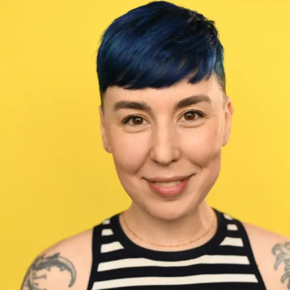 Person with short blue hair, wearing a black and white striped sleeveless top, smiles in front of a solid yellow background. Tattoos are visible on both shoulders.