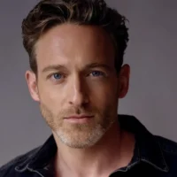 A man with short, wavy brown hair, light beard, and blue eyes looks at the camera. He is wearing a dark shirt and is against a plain, neutral background.