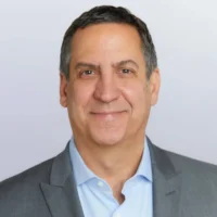 A middle-aged man with short dark hair wearing a light blue shirt and grey suit jacket, smiling at the camera against a plain background.