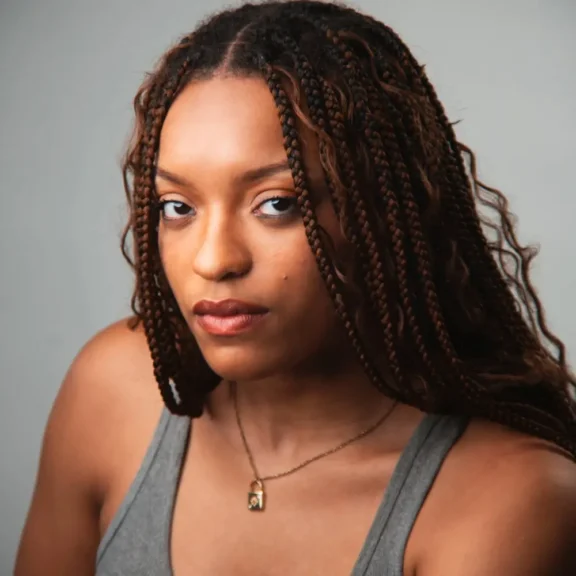 A woman with long, braided hair wearing a gray tank top and a necklace looks at the camera against a plain background.
