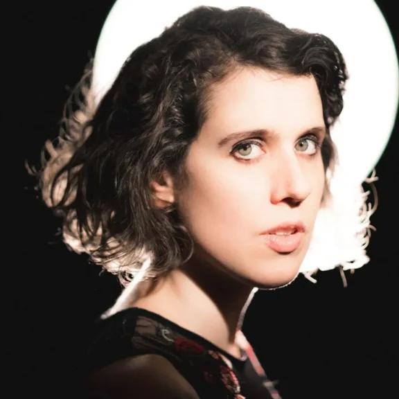 A woman with short, wavy dark hair is shown in front of a bright circular light, looking at the camera with a serious expression. The background is dark.