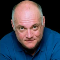 A bald man with light skin and green eyes wearing a blue collared shirt looks directly at the camera against a black background.