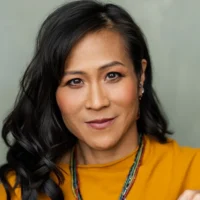 Woman with long black hair and a yellow top looks directly at the camera, wearing layered necklaces and subtle makeup, against a neutral background.