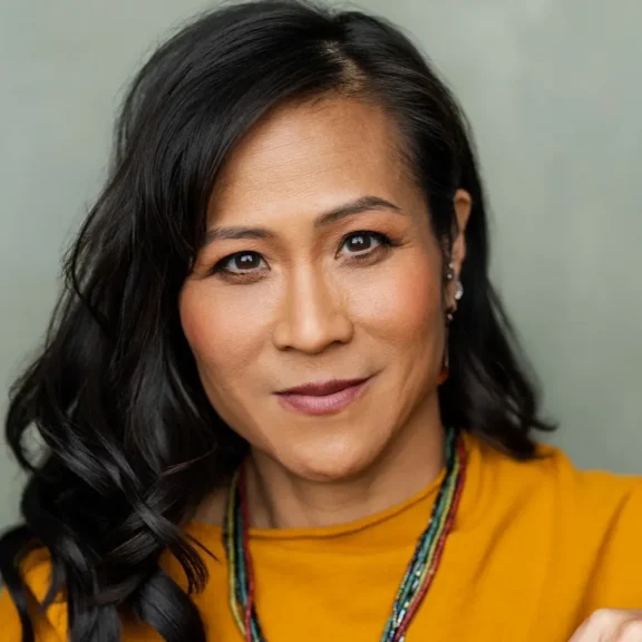 Woman with long black hair and a yellow top looks directly at the camera, wearing layered necklaces and subtle makeup, against a neutral background.