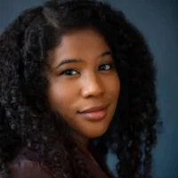 A woman with long, curly dark hair and a brown jacket looks at the camera against a blue background.