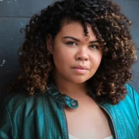 Woman with curly hair wearing a teal leather jacket looks directly at the camera against a dark, textured background.