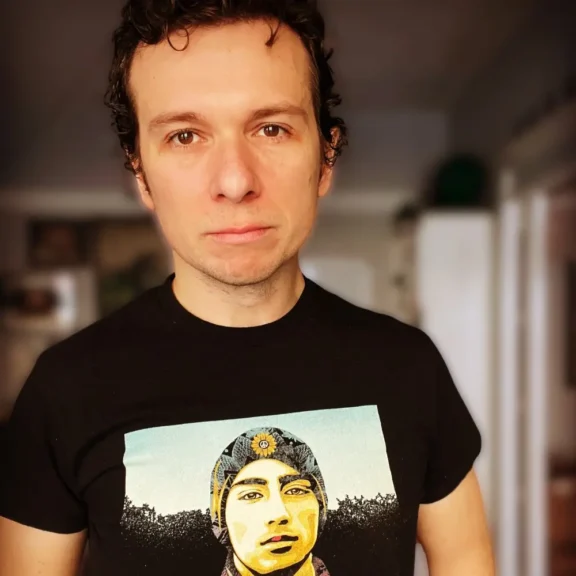 A man with short, curly dark hair wears a black t-shirt featuring an illustrated portrait of a person in a knit hat. He stands indoors and looks directly at the camera.