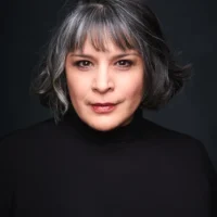 Woman with short, wavy salt-and-pepper hair wearing a black turtleneck, looking directly at the camera against a dark background.