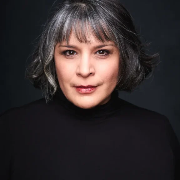 Woman with short, wavy salt-and-pepper hair wearing a black turtleneck, looking directly at the camera against a dark background.
