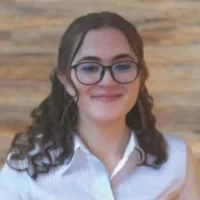 A person with long brown curly hair and glasses smiles, wearing a white collared shirt, standing in front of a wooden background.