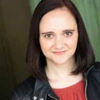 Woman with straight dark hair wearing a black leather jacket over a red top, smiling slightly, standing in front of a green background.