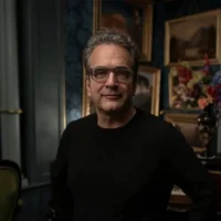 A middle-aged man with glasses and gray hair stands indoors, wearing a black shirt. Framed paintings and flowers are visible in the dimly lit background.
