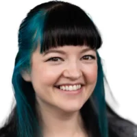 A woman with teal and black hair, blunt bangs, and fair skin is smiling, facing the camera against a plain white background.
