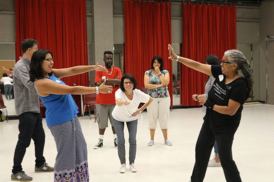 A group of five people in a rehearsal room with red curtains. The group is acting out an exercise during a workshop.