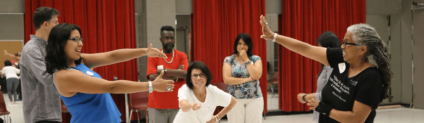 A group of five people in a rehearsal room with red curtains. The group is acting out an exercise during a workshop.