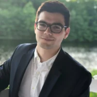 A person wearing glasses and a dark blazer over a white shirt sits outdoors in front of a body of water and trees.