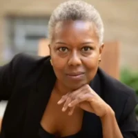 A woman with short gray hair wearing a black blazer, resting her chin on her hand.