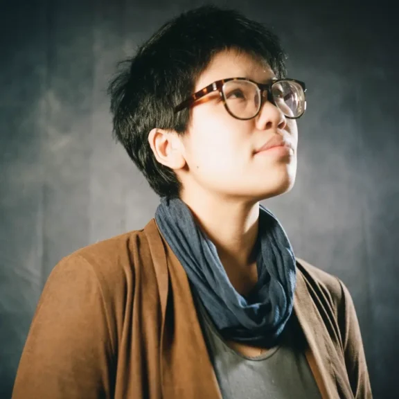Person with short dark hair and glasses, wearing a brown blazer and blue scarf, looks upward against a dark, softly lit background.