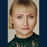 A person with short blonde hair and blue eyes is wearing a green lace top, looking directly at the camera with a neutral expression.