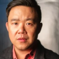 A man with short dark hair, wearing a dark jacket over a red collared shirt, looks directly at the camera against a neutral background.