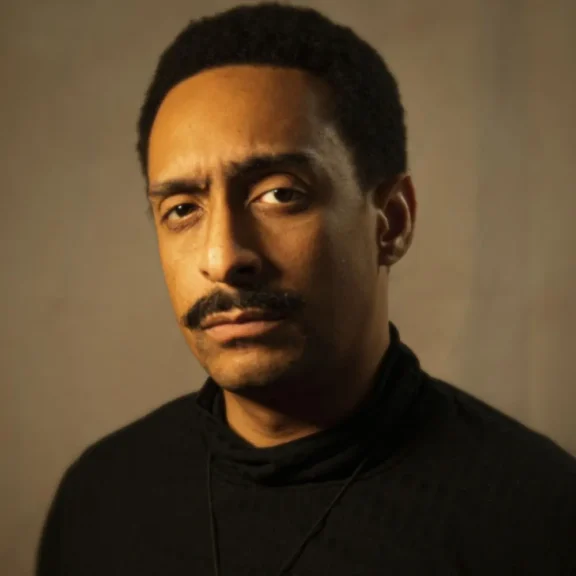A man with short curly hair and a mustache wears a black turtleneck and looks directly at the camera with a neutral expression against a plain background.