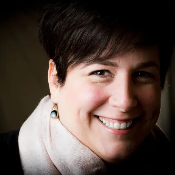 A woman with short dark hair, wearing a light scarf and a single earring, smiles at the camera against a dark background.