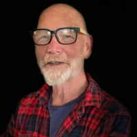 Bald man with a white beard wearing large black glasses, a red plaid shirt, and a dark T-shirt, standing against a plain black background.