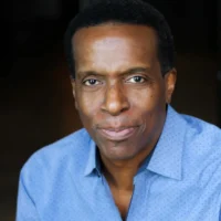 A middle-aged man with short dark hair wearing a light blue, button-up shirt, looking directly at the camera with a neutral expression.