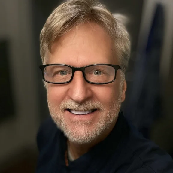 A middle-aged man with light hair and a beard, wearing glasses and a dark shirt, smiles at the camera indoors.