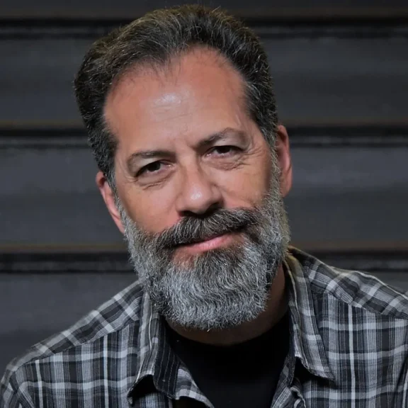 A middle-aged man with gray hair and a full beard, wearing a plaid shirt, looks at the camera with a slight smile. Dark, blurred horizontal stripes are in the background.