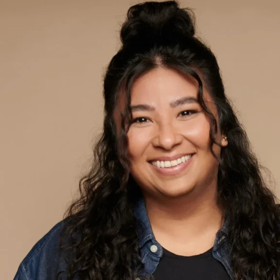 A woman with long curly hair styled half up, wearing a denim jacket over a black top, smiles at the camera against a plain beige background.