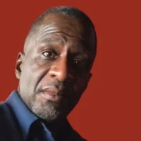 A man with short hair and a trimmed beard wearing a dark blue shirt poses against a solid red background.