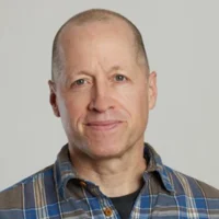 A middle-aged man with a shaved head wearing a blue plaid shirt, standing against a plain light gray background.