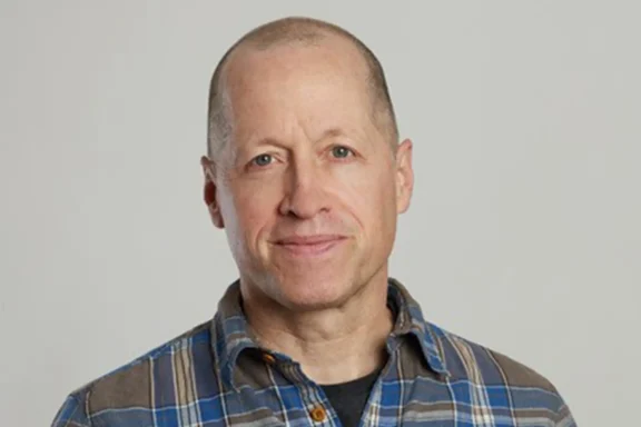A middle-aged man with a shaved head wearing a blue plaid shirt, standing against a plain light gray background.