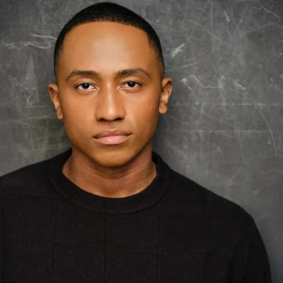 A man with closely cropped hair and a neutral expression stands in front of a textured gray background, wearing a black sweater.