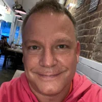 A man wearing a pink hoodie smiles at the camera while sitting in a restaurant with brick walls and hanging lights.