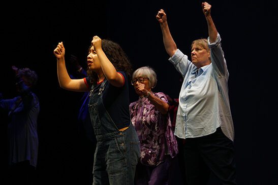 Three people on stage looking out into the audience, with their hands raised as if they were holder banners in front of them.