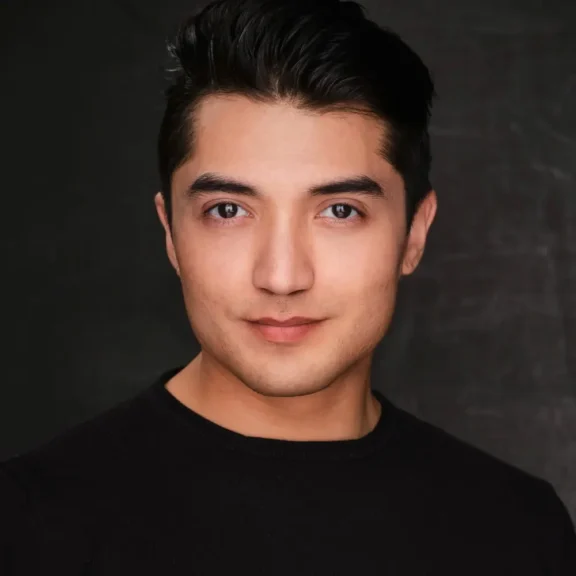 Young man with short dark hair and a clean-shaven face, wearing a black crew-neck shirt, poses against a dark, plain background.