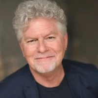 Smiling older man with curly gray hair and beard, wearing a dark blazer and black shirt, posed against a neutral blurred background.